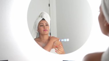 Havlu giymiş bir kadın banyoda vücut kremi sürüyor. Duştan sonra doğal nemlendirici tedaviyle kişisel cilt bakımını sergiliyor.