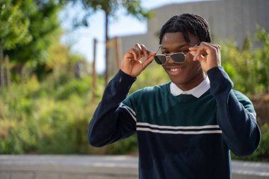 Parkta dikilirken güneş gözlüğünü ayarlayan gülümseyen genç bir öğrencinin portresi.