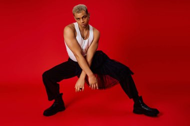 Genç bir adam atlet bluzuna ve siyah pantolona güveniyor, canlı kırmızı bir arkaplanda poz veriyor..