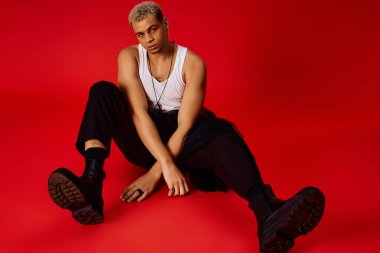 Siyah pantolonlu, kolsuz tişörtlü genç bir adam kırmızı bir stüdyoda şık poz veriyor..