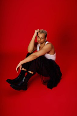 Saçları boyalı genç adam kırmızı bir stüdyoda şık poz veriyor, kendine güveni ve modası var..