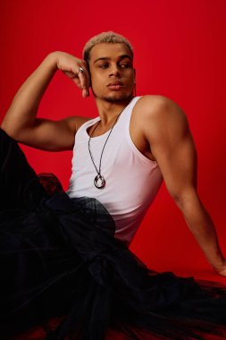 Tarz sahibi genç bir adam, kırmızı bir fonda siyah pantolon ve kolsuz bir bluzla rahatça poz verir..