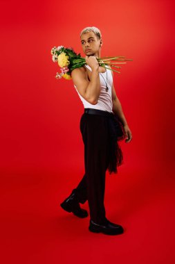 Genç bir adam eşsiz bir moda seçimi sergiliyor. Parlak kırmızı bir stüdyoda çiçeklerle zarif bir poz veriyor..
