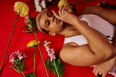 Tarz sahibi genç bir adam kırmızı bir arka planda renkli çiçeklerle çevrili boyalı saçlarıyla poz verir..