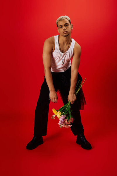 Young man with dyed hair striking a pose while holding vibrant flowers. The background is red.