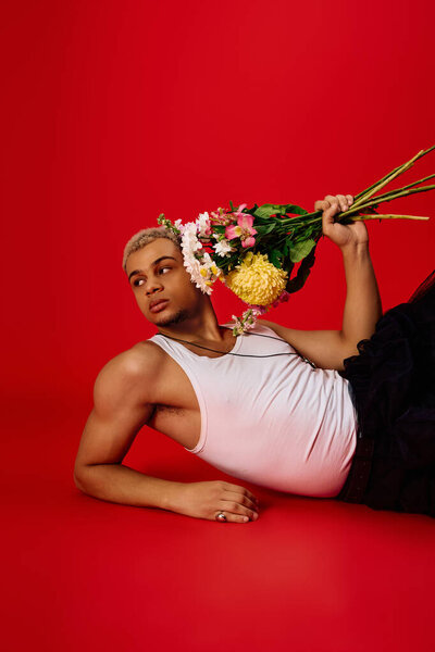 A young man in a tank top with dyed hair poses with flowers against a red background.