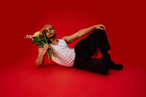A young African American man poses stylishly with flowers in a striking red studio setting.