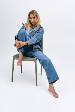 Sarışın kadın stüdyo ortamında bir sandalyede otururken şık bir şekilde kot giyip poz veriyor..