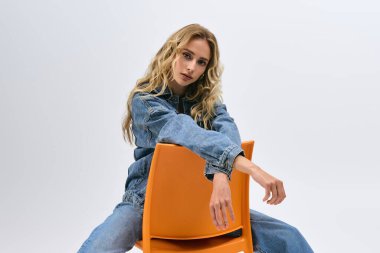 Sarışın kadın şık bir şekilde turuncu bir sandalyeye uzanmış modaya uygun kot giyim tarzını sergiliyor..