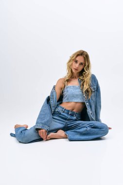 Sarışın, kendine güvenen genç bir kadın zarif bir şekilde otururken şık bir kot elbise sergiliyor..