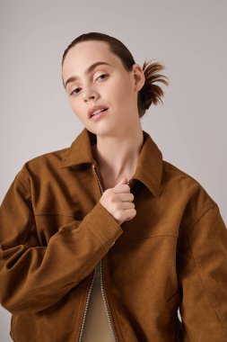 Kahverengi ceketli genç bir kadın tarafsız bir arkaplana karşı güvenle poz veriyor..