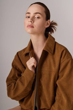 Kendine güvenen bir kadın, minimalist bir arkaplan karşısında şık kahverengi bir ceket mankenliği yapıyor..