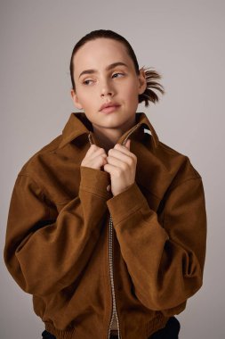 Genç bir kadın, tarafsız bir arkaplan karşısında modaya uygun kahverengi bir ceketle çağdaş bir tarz sergiliyor..