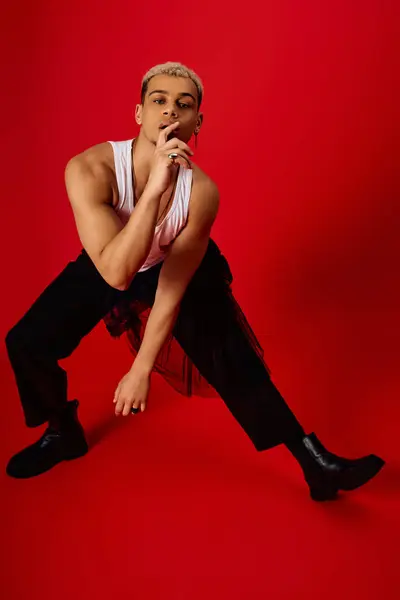 A young man poses stylishly in a red studio wearing a tank top and black pants, showcasing fashion. — Stock Photo