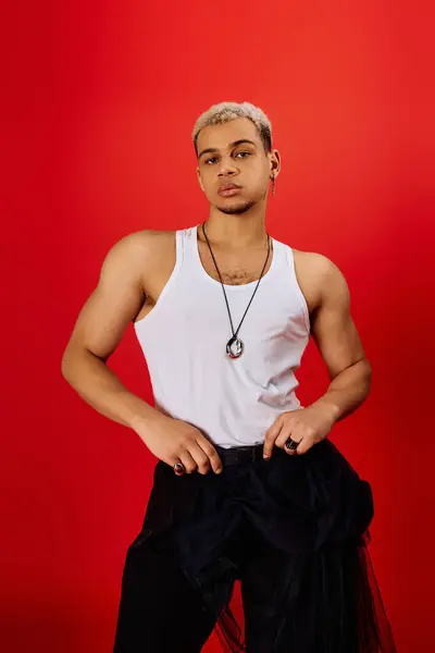 A young man with dyed hair poses confidently in a tank top and black pants against red. — Stock Photo