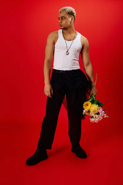 A young African American man poses stylishly in a red studio, holding colorful flowers in hand. — Stock Photo