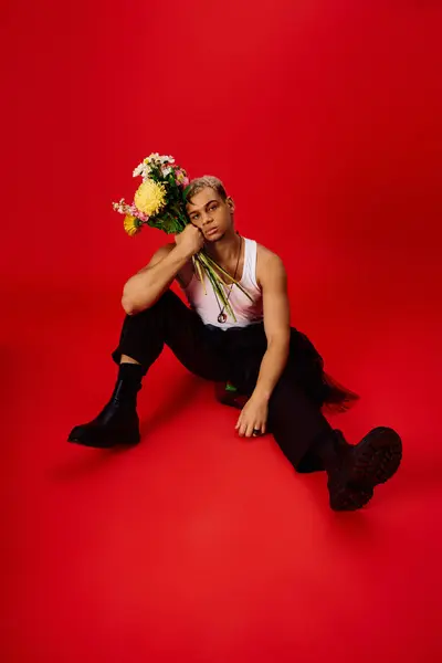 A fashionable young African American man poses gracefully in a red studio, holding flowers. — Stock Photo