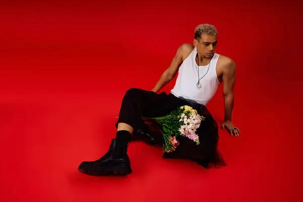 A young African American man poses stylishly in a red studio, holding vibrant flowers in his lap. — Stock Photo