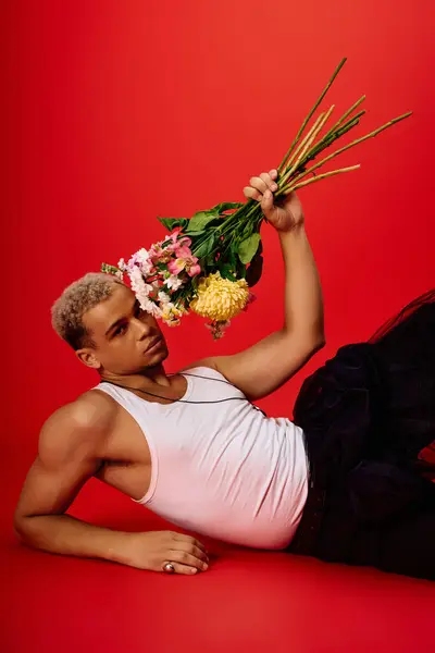Young man poses stylishly on a red backdrop, holding vibrant flowers while dressed in a tank top. — Stock Photo