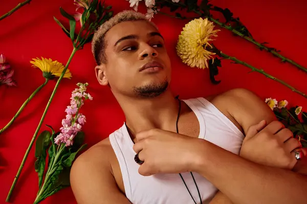 A stylish young man with dyed hair poses in a trendy tank top, surrounded by colorful flowers. — Stock Photo