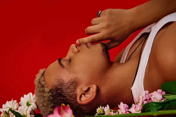 A stylish young man with dyed hair reclines gracefully among flowers in a red studio setting. — Stock Photo