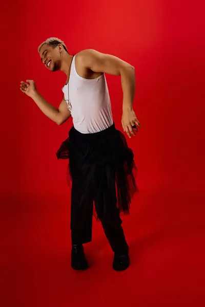 A young man with dyed hair poses stylishly in a red studio, wearing a tank top and black pants. — Stock Photo