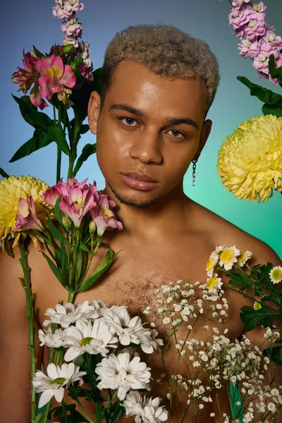 A young, shirtless man with dyed hair poses confidently among colorful flowers in a studio. — стокове фото