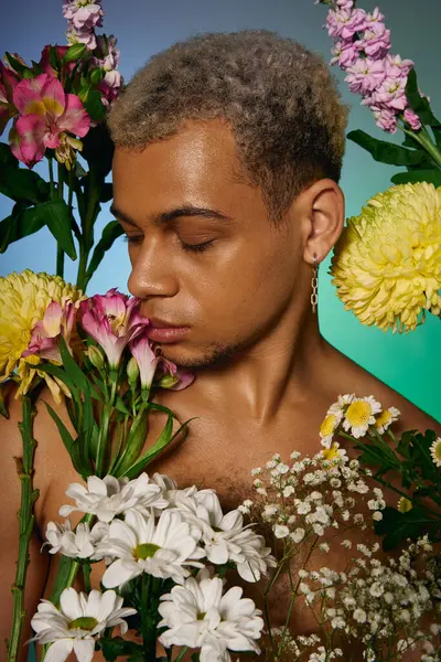 Muscular young man with dyed hair poses serenely among colorful flowers in a studio. — Stock Photo