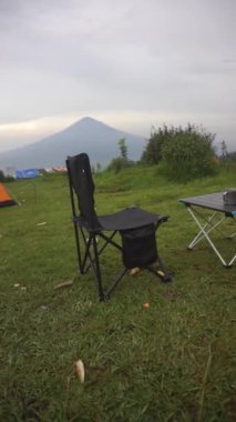 Açık tepelerdeki yeşil çimlerin üzerinde katlanan bir masanın olduğu bir fincan kahvenin dikey video görüntüsü ve siyah katlanır bir sandalye.