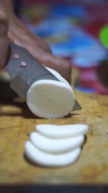 Bir adamın elleri, temiz ve beyaz gözüken bir hayatta kalma bıçağıyla manyok doğrayıp soyar..