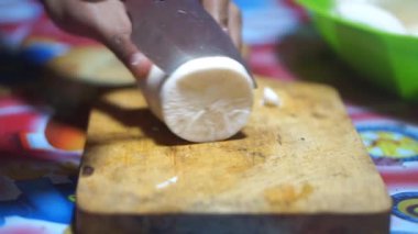 Bir adamın elleri, temiz ve beyaz gözüken bir hayatta kalma bıçağıyla manyok doğrayıp soyar..