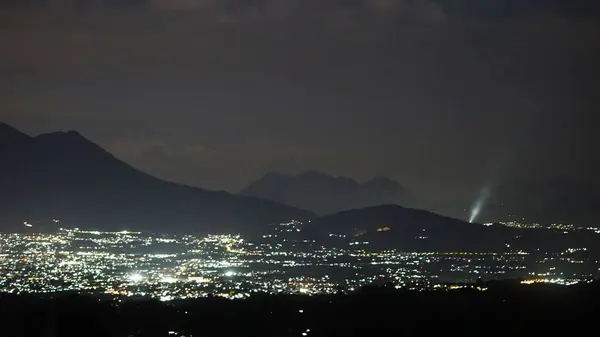 Dağlık bir ufkun altında parlayan şehir ışıkları, karanlık çöktükten sonra doğa ve şehir yaşamı arasındaki zıtlığı vurguluyor.