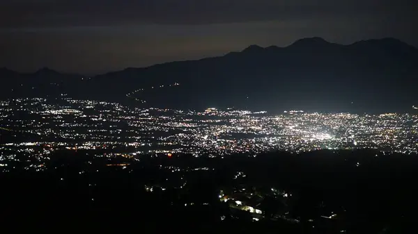 Dağlık bir ufkun altında parlayan şehir ışıkları, karanlık çöktükten sonra doğa ve şehir yaşamı arasındaki zıtlığı vurguluyor.
