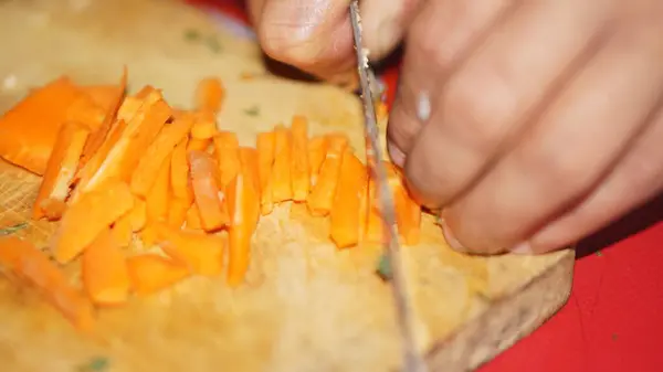 Mutfak bıçağıyla ahşap bir kesme tahtasının üzerinde taze havuçları kesen ellerin yakın çekimi..