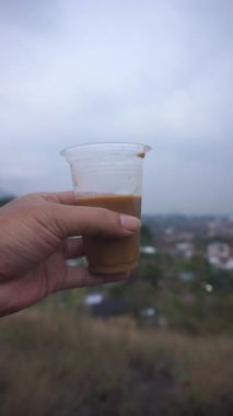 Elinde plastik bir bardak buzlu kahve sütü ve manzaralı şehir manzarası var..