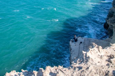 An angler on the rocks of the cliff fishing in the breaking waves. Coastal landscape of the archaeological site on the coast of Roca Vecchia, Lecce, Italy.