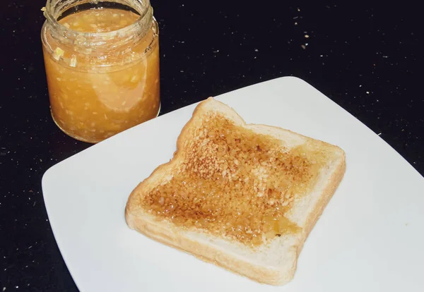 Toast spread with orange marmalade in Madrid, Spain. Toast presented on a white plate next to the glass jar of homemade jam.