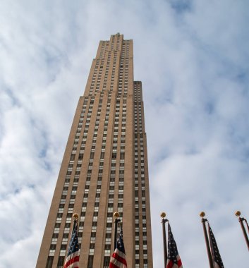 New York şehrinin ufuk çizgisinin altından bir manzara, arkasında bulutlu bir gökyüzü. Üssünde Amerikan bayrakları bulunan Uluslararası Bina (Rockefeller Center) manzarası.
