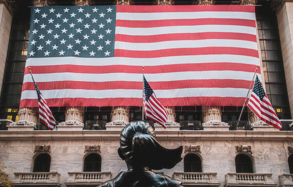 Статуя Бесстрашной Девушки перед Нью-Йоркской фондовой биржей, Нью-Йорк, США. Американский флаг на фасаде фондовой биржи. Уолл-стрит, ноябрь 2019.