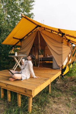 Genç kadın, kamp ateşinin yanındaki koltukta rahatça otururken dizüstü bilgisayarda çalışıyor. Çadırla seyahat ediyor. Uzak çalışma kavramı ve doğaya kaçmak
