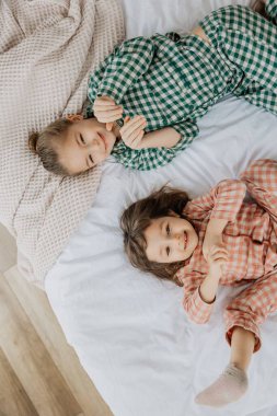 Yatakta el ele uzanmış, kameraya bakıp gülümseyen neşeli küçük kız kardeşlerin üst manzara portresi. Çocuk ilişkileri ve değerleri