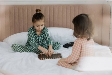 Aynı pijamaları giyen iki kız, sabahları uyuduktan sonra ailelerinin yatak odasındaki yatakta masa oyunu oynarlar.