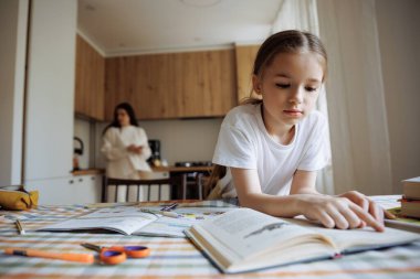 6 yaşında bir kız çocuk kitap okuyor ve annesinin mutfakta yemek pişirme geçmişine karşı ödev yapıyor.