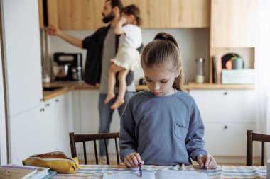 6 yaşındaki kızının ödevlerini kontrol eden bir baba mutfak masasında oturan ve mutfakta yemek pişiren babasının önünde ödevlerini yapan bir kız.