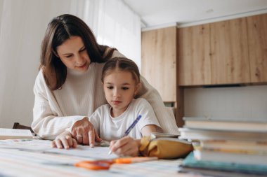 Annesi ile yemek masasında ödev yapan genç bir kız modern bir mutfakta arka planda çalışıyor..