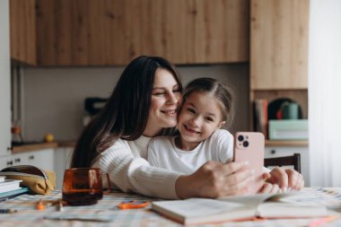 Mutlu anne-kız telefonda, evde, mutfak masasında fotoğraf çekiyor.