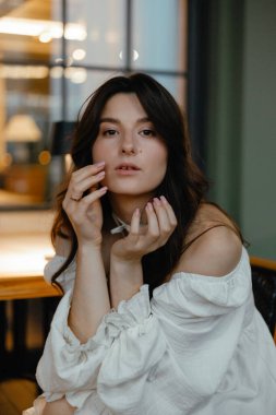 Açık tenli, beyaz bir elbise giyen genç ve seksi bir esmer, akşam bir sokak restoranında masada otururken fotoğraf için poz veriyor.