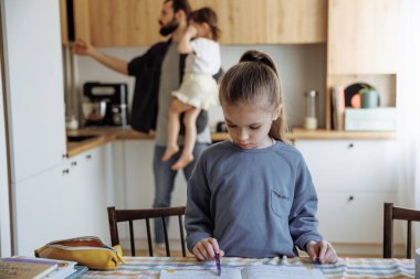 6 yaşındaki kızının ödevlerini kontrol eden bir baba mutfak masasında oturan ve mutfakta yemek pişiren babasının önünde ödevlerini yapan bir kız.