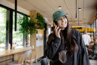 Elinde telefonuyla gülümseyen çekici bir kız bir kahve dükkanının önünde durmuş arkadaşlarını ya da siparişini bekliyor ve telefonu kullanıyor. 