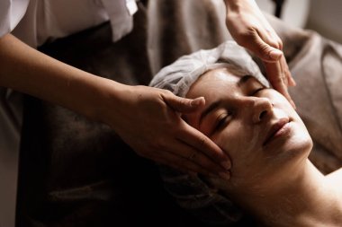 Güzel bir kadının yakın plan portresi, yan ışığı olan bir güzellik merkezinde nemlendirici ile yüz masajı yaptırıyor. Cilt bakımı için doğal kozmetik ürünleri. Sağlık hizmetleri, güzellik